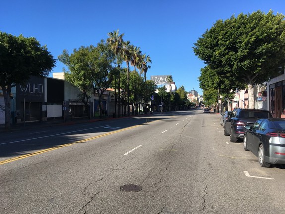 Zu sehen ist eine leere Straße, an den Seiten stehen Bäume und Palmen und ein paar Autos parken am Straßenrand